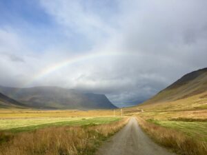 A long dirt road through an endless grassfield between two ridges with a rainbow spanning from ridge to ridge. Happiness is about the and.