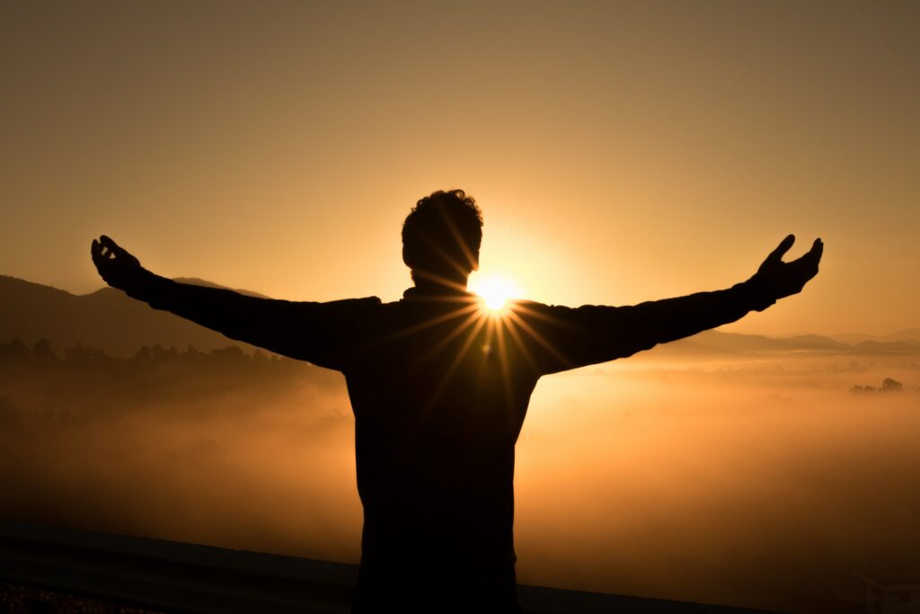 A man standing on a mountain top at sunrise with arms outstretched; he is letting go and allowing