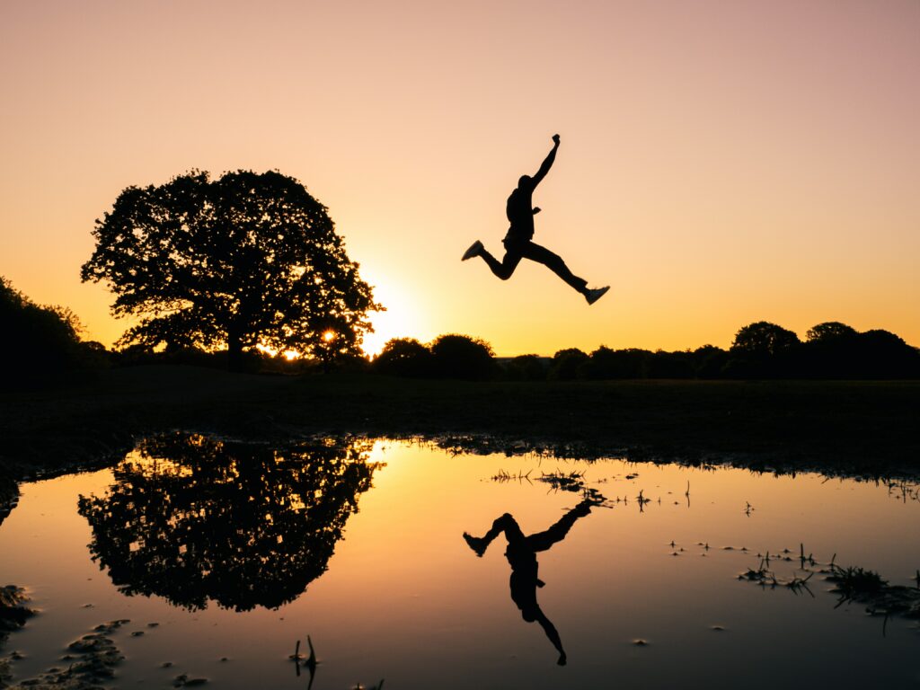 A man in silhouette taking a huge leap over a wide water way reflecting both the man and the setting sun's magenta sky. He is not going to wait any longer.