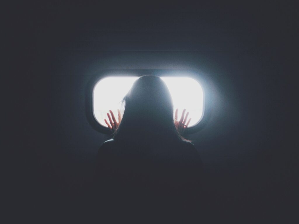 A woman in the dark looking out  to the light through a small horizontally oblong window. Her hands are pressed against the window. What is she waiting for?