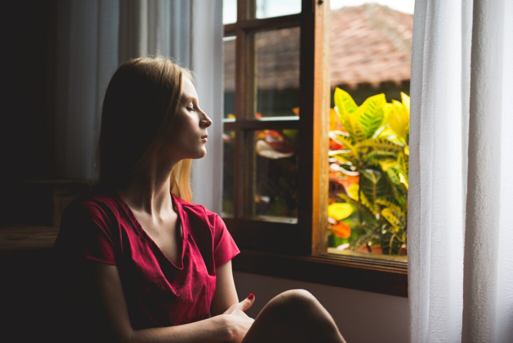 A woman sits by an open window appearing to turn within to find the answers she is waiting for. 