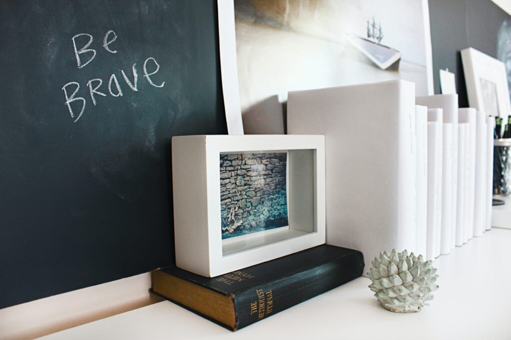 A message of Be Brave written on a chalk board next to a stack of books of yet to be written stories. 
