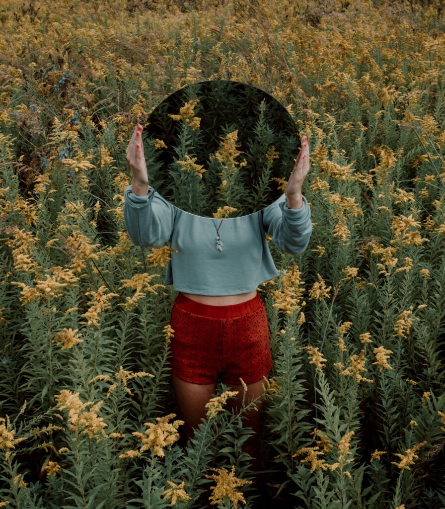 A woman standing in a field of Golden Rod holding a mirror to her face that the tells the story of her as Golden Rod.