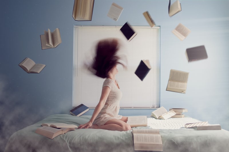 A woman kneeling on a bed flipping her hair in anger and frustration with books surrounding her on the bed and flying through the air.