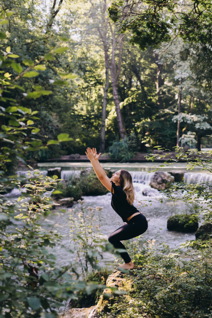 A women next to a rambling river in the woods in the yoga position “chair” as she physically moves into her edge 