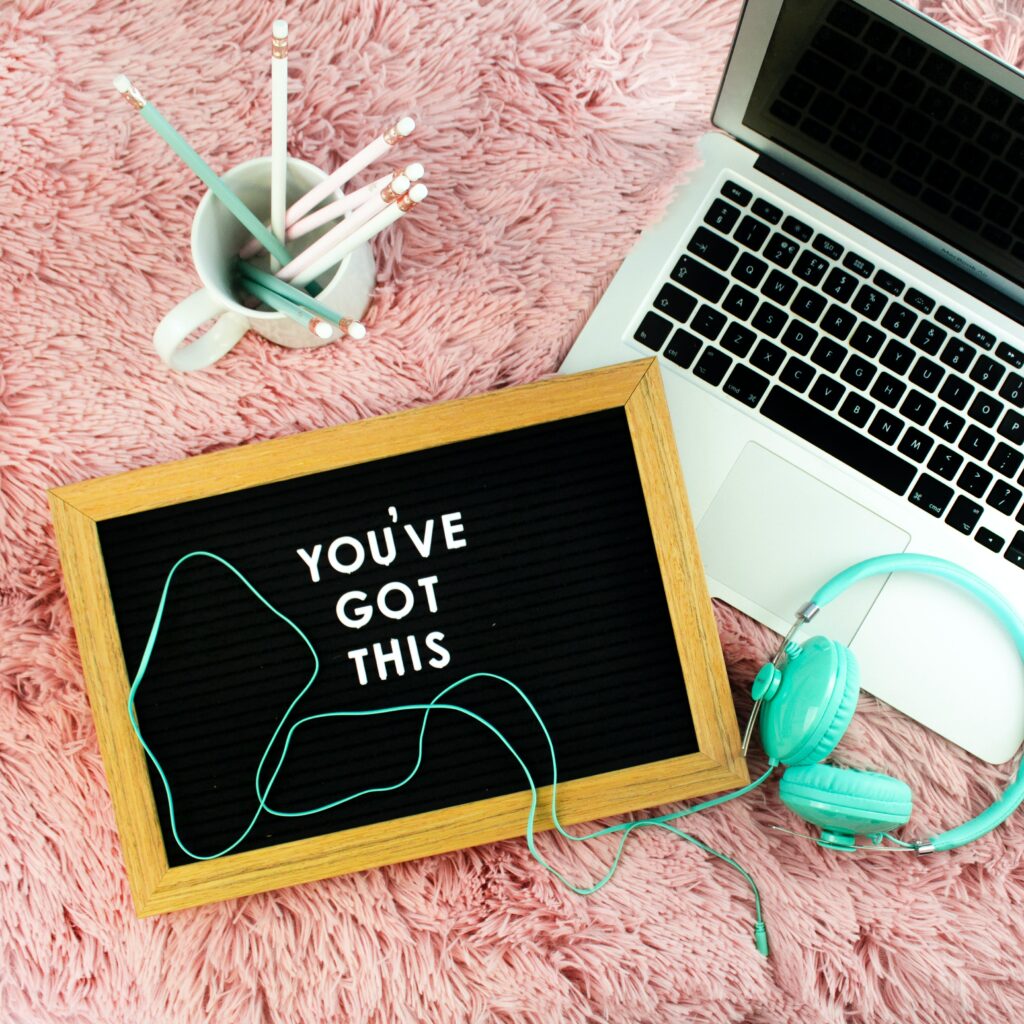 I laptop, headphones, cup of pens and a sign that says "you've got this" on pink shaggy background
