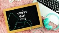 I laptop, headphones, cup of pens and a sign that says "you've got this" on pink shaggy background