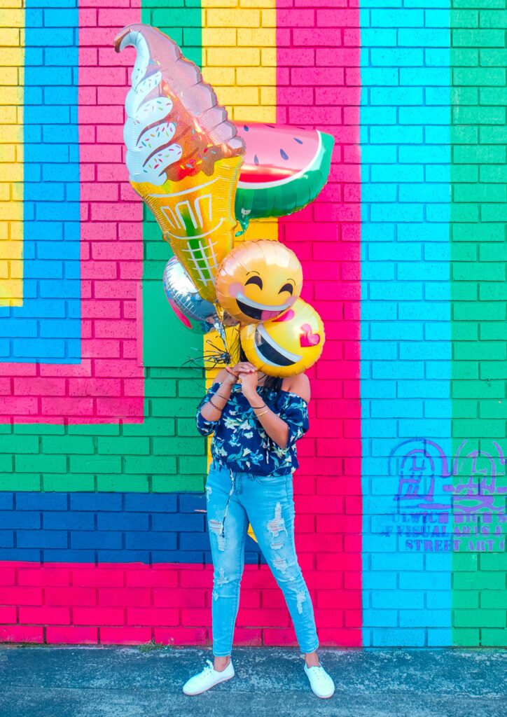 A woman behind a large, colorful geometric design on a brick wall playing and celebrating with big balloons in front of her face.
