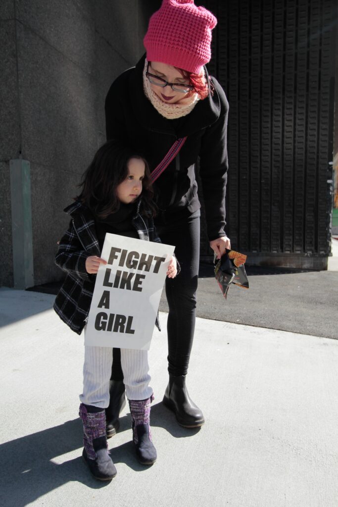Woman leaning over girl holding sign that reads “fight like a girl”