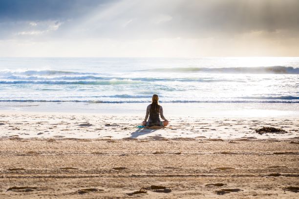 A woman sits with back to camera folded legs, yoga style facing a irridescent sea